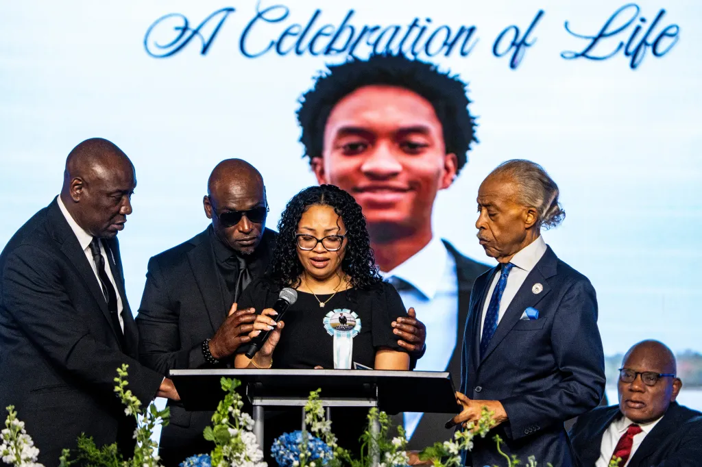 Christine Wonsley speaks at a funeral for Nolan Wells, consoled by Ben Crump, Elmore Wonsley, and Rev. Al Sharpton.