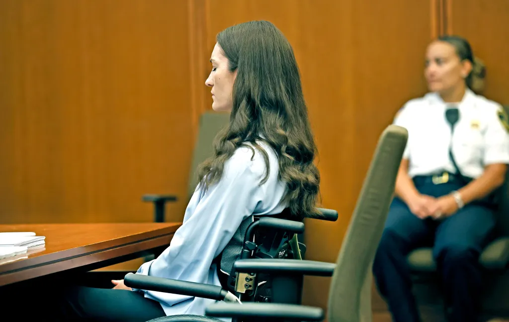 Lindsay Clancy, in a wheelchair, in court, with an officer nearby.