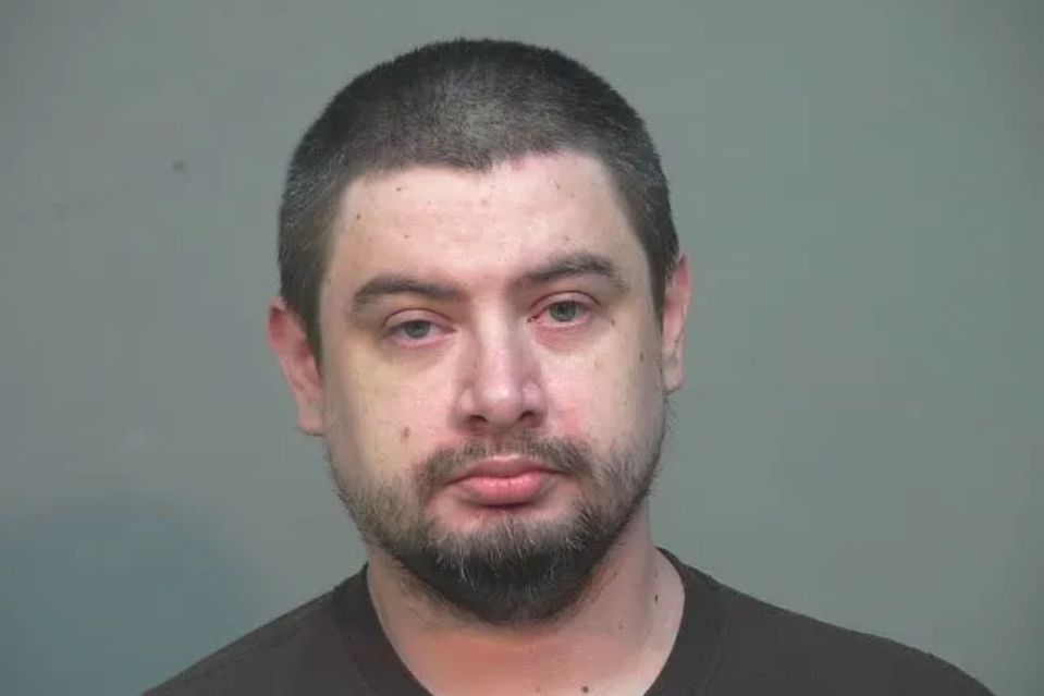 An image collage containing 1 images, Image 1 shows Mugshot of a man with short dark hair and a beard, wearing a dark t-shirt, looking directly at the camera