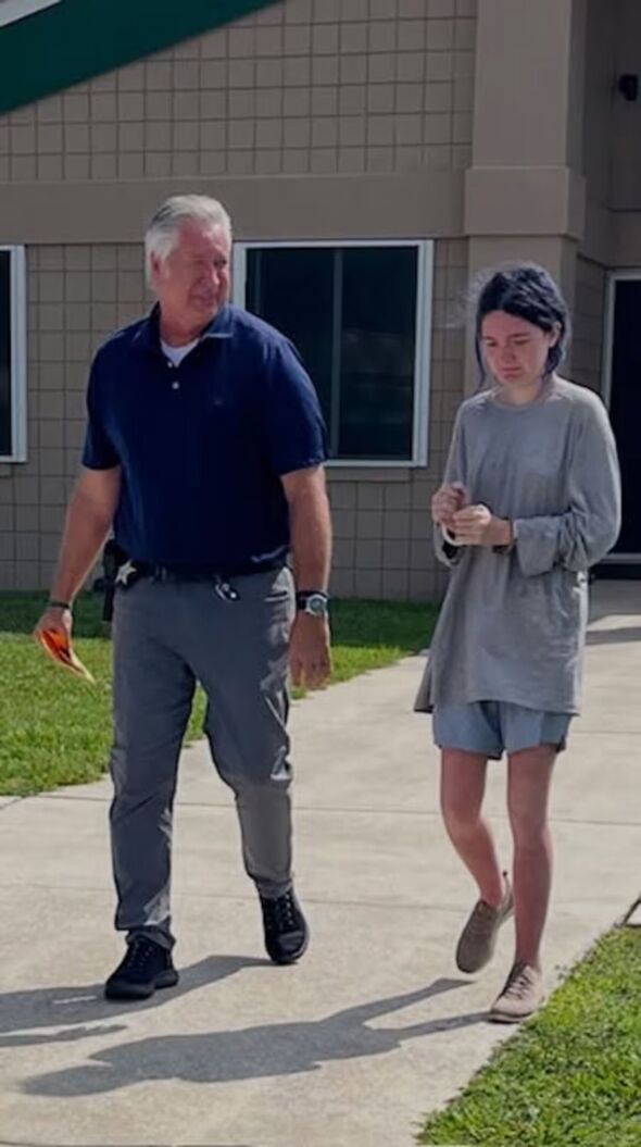 A hunched woman walks alongside a law enforcement officer