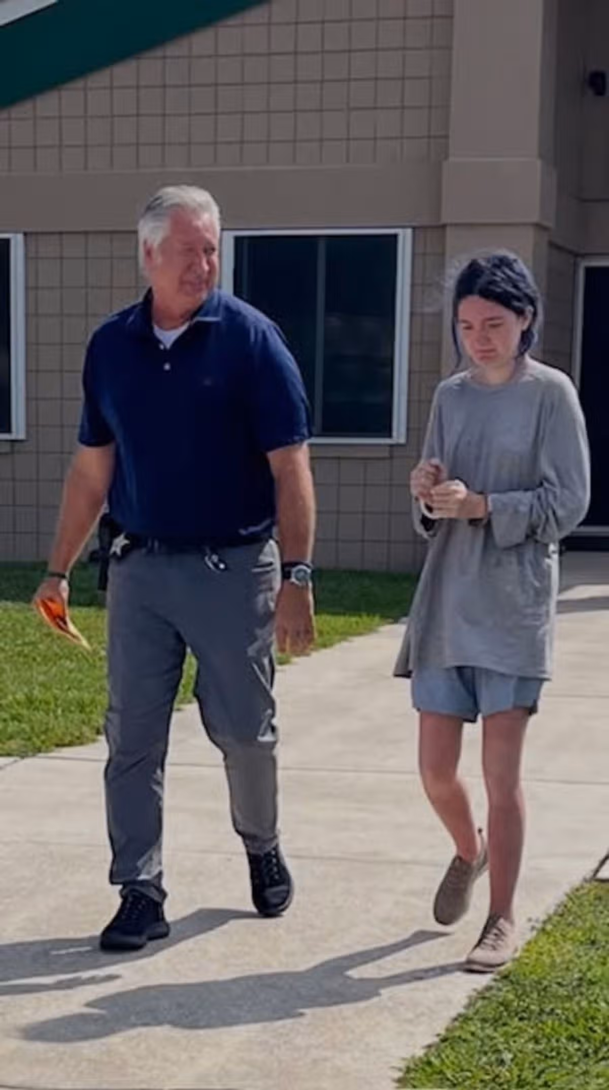 A hunched woman walks alongside a law enforcement officer