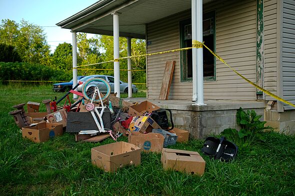 Various boxes, items, and a bicycle are scattered on the lawn in front of a house. The property is cordoned off with yellow tape