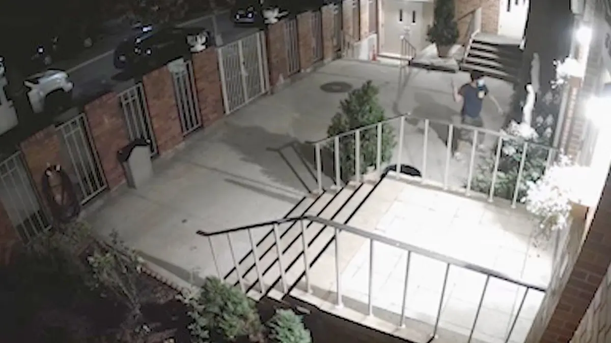 Masked man in dark shirt jumps over fence into church courtyard.
