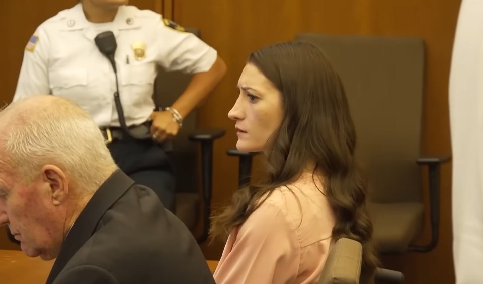 A woman with long dark hair sits in a courtroom, looking to the side, with a police officer standing behind her.