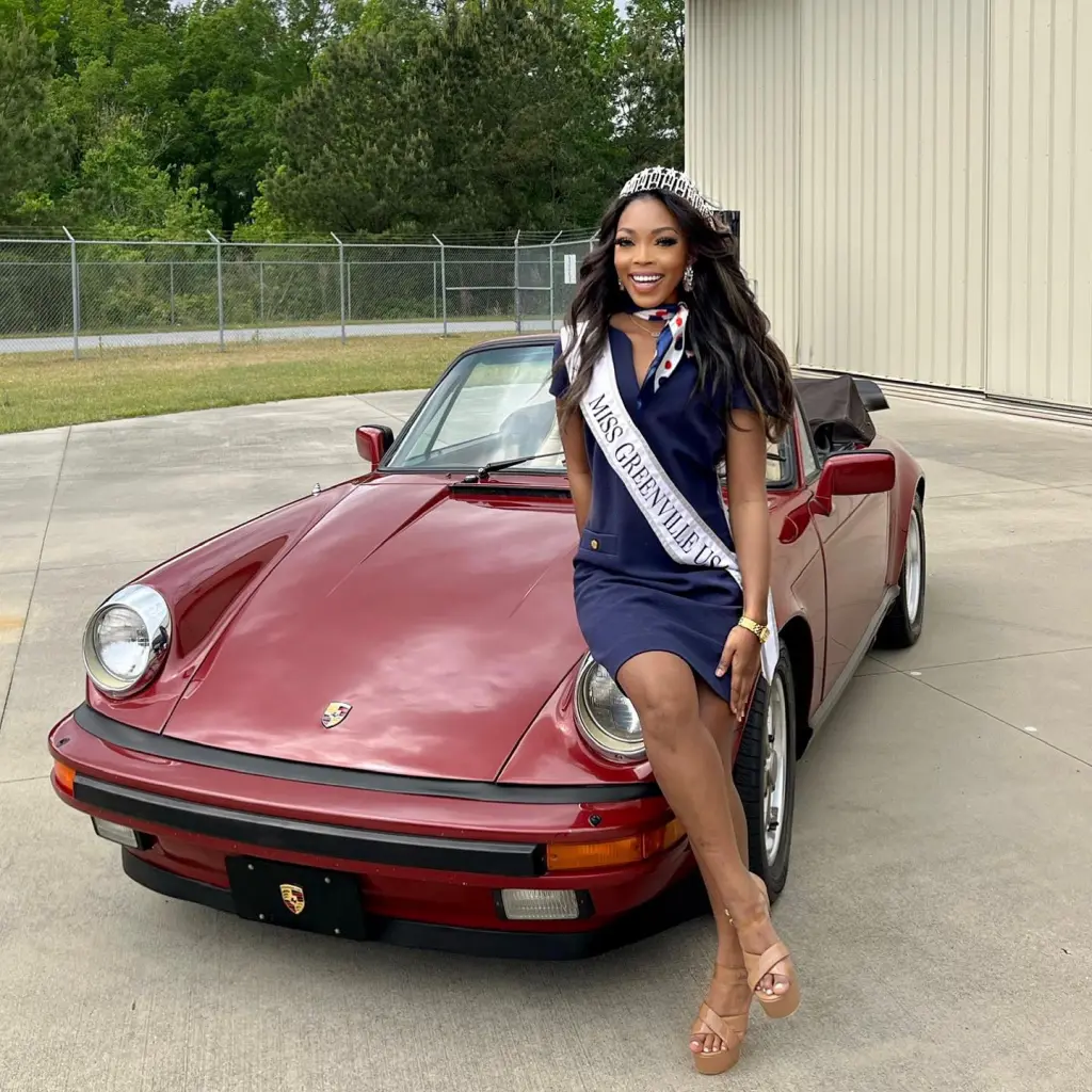 Alexis Yearde, wearing a "Miss Greenville US" sash, a tiara, and a navy dress, sits on the hood of a red convertible.