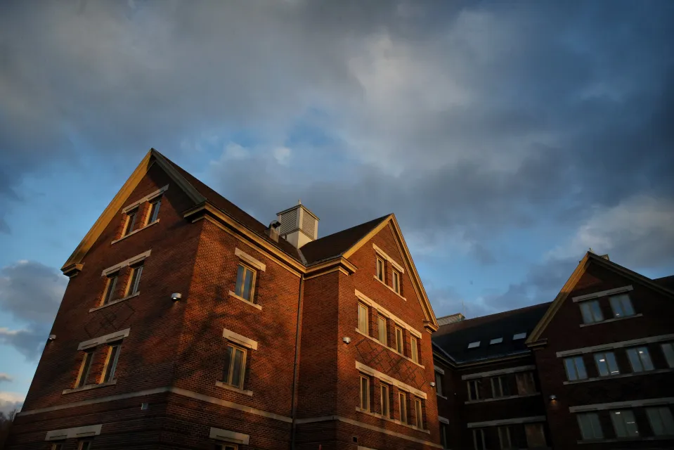 A low-angle photo of a brick building with several windows, against a partly cloudy sky.