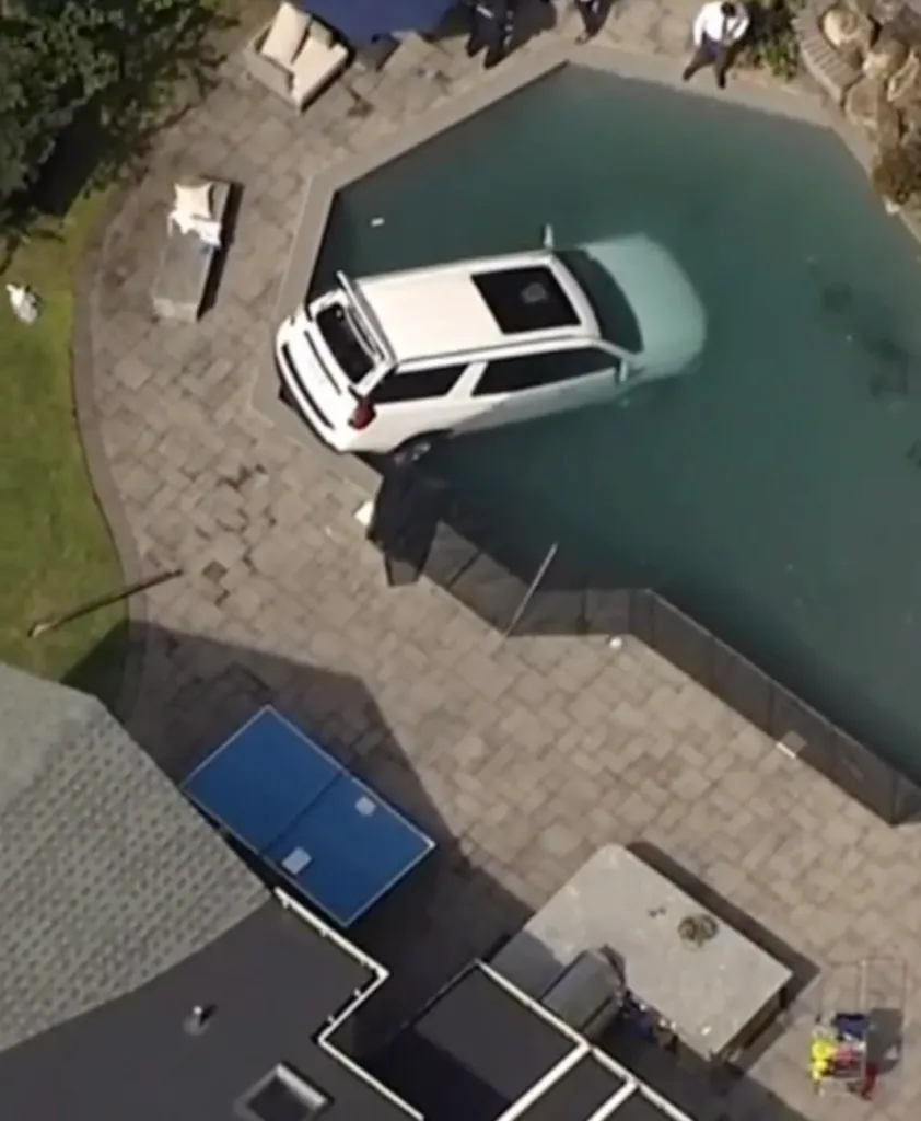 An aerial view shows a white SUV partially submerged in a swimming pool next to a patio.