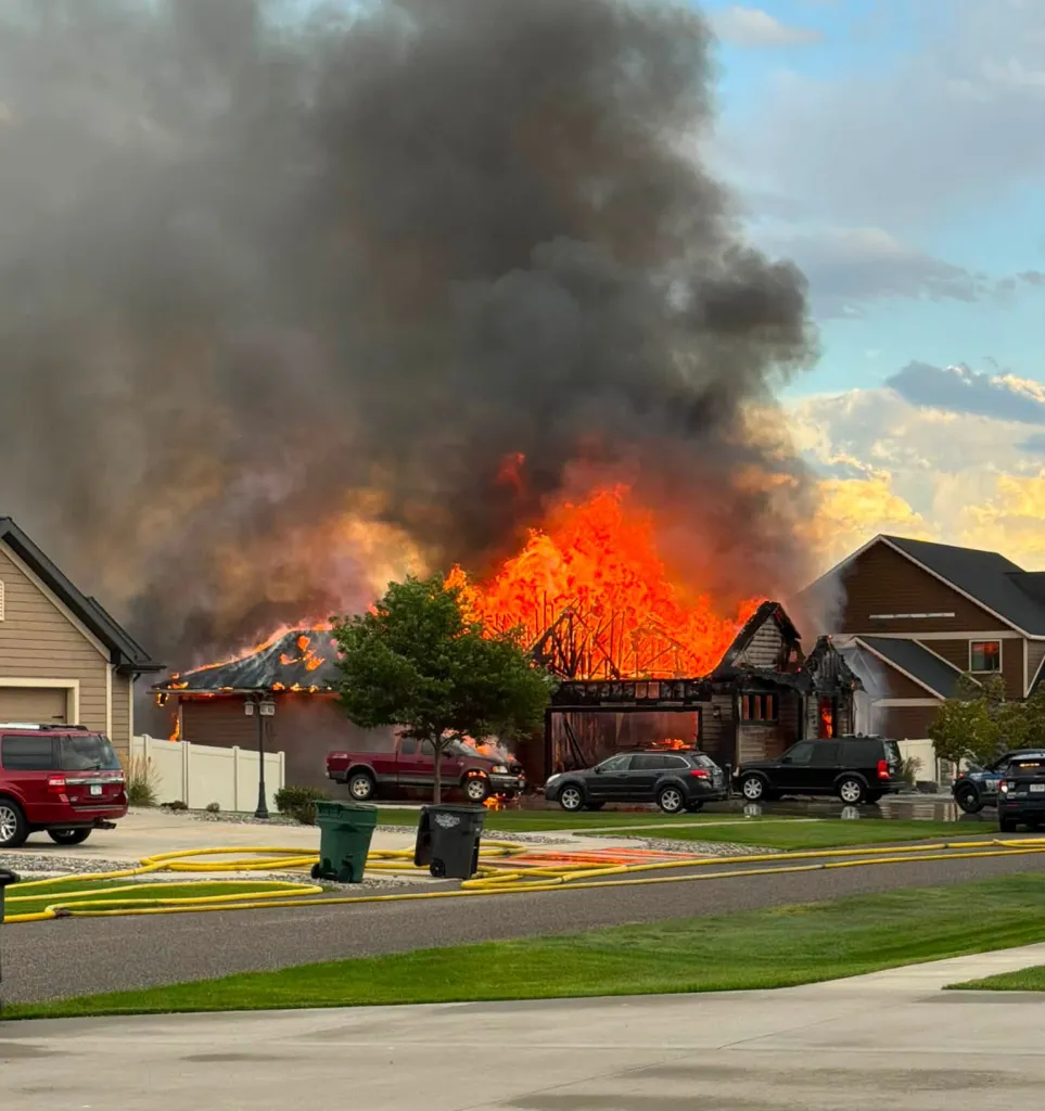 A house engulfed in large flames and thick smoke with several cars parked outside.