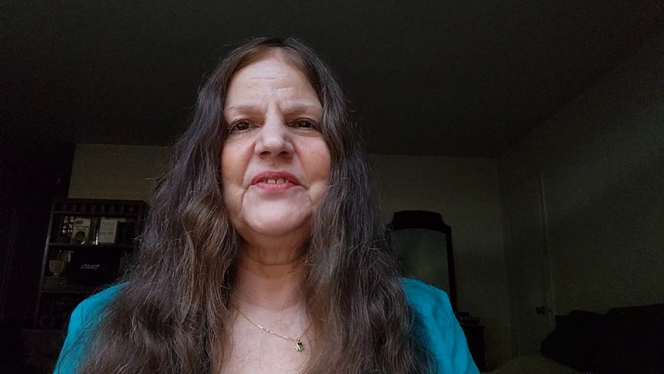 A woman with long brown hair wearing a teal top and a gold necklace smiles at the camera.