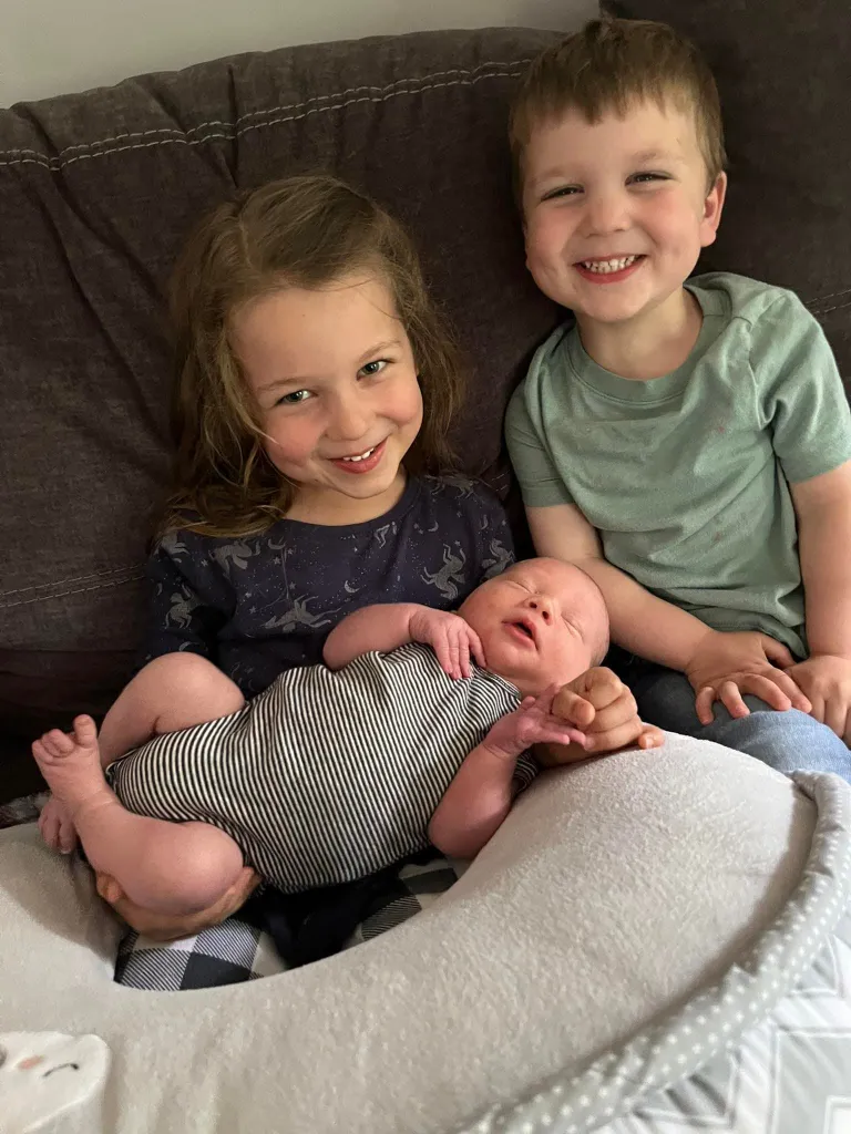 Cora, Dawson, and their baby sibling smiling while sitting on a couch.