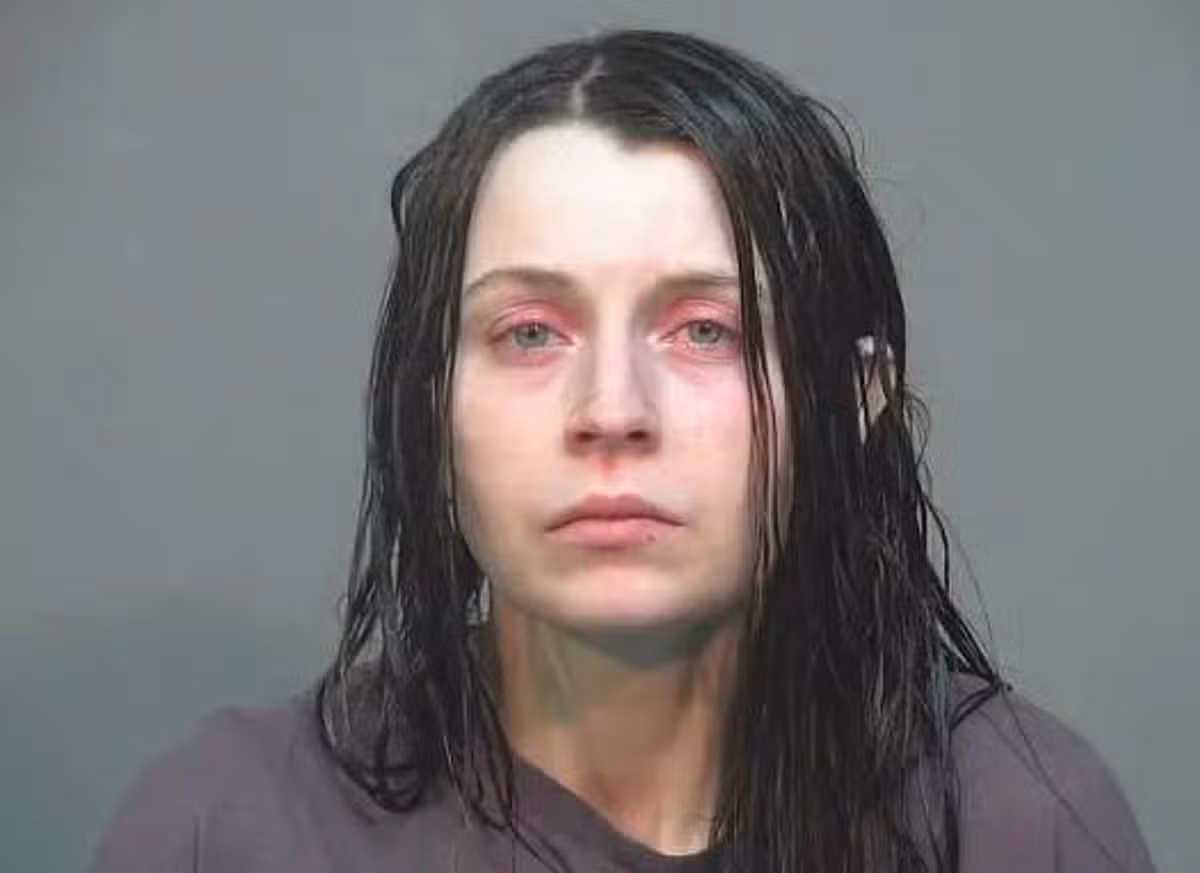 An individual with long, wet black hair and a somber expression stands against a plain gray background. The person appears to be in a formal setting, likely for a photograph taken for official purposes.