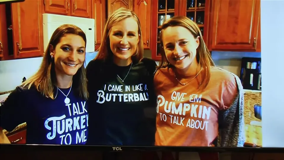 Three women wearing Thanksgiving-themed shirts in a kitchen.
