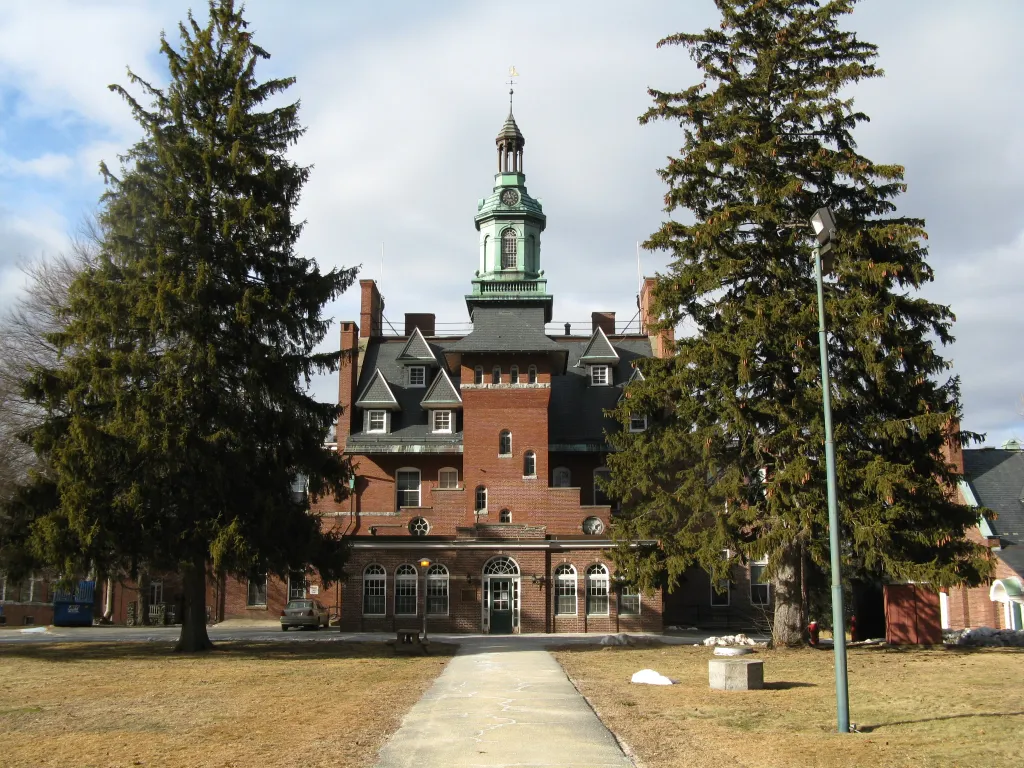 Tewksbury State Hospital, a large brick building with a green clock tower, with a pathway leading to the entrance and two large evergreen trees framing the building.