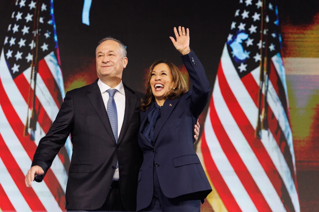 Vice President Kamala Harris and her husband Doug Emhoff stand together, smiling, with American flags behind them at the DNC Convention.