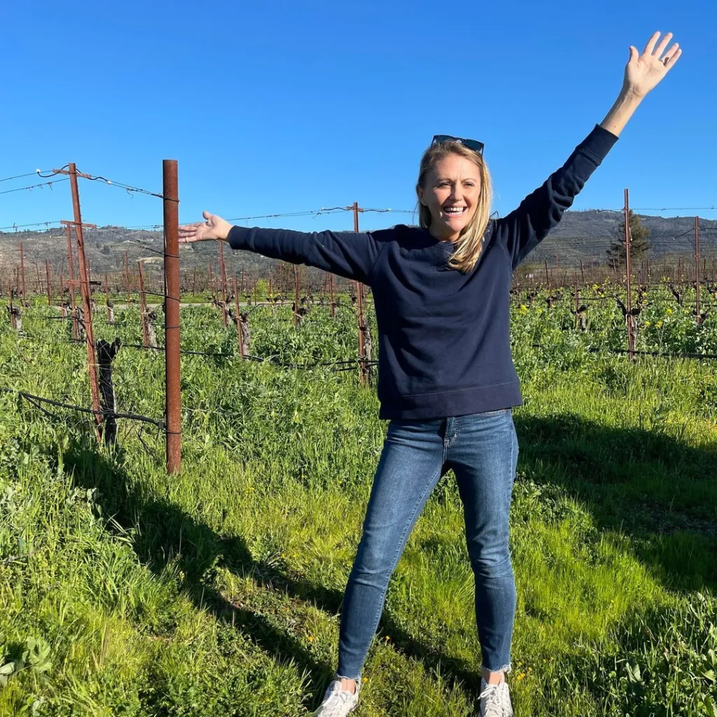 Brandee Mulligan stands in a vineyard with her arms raised.