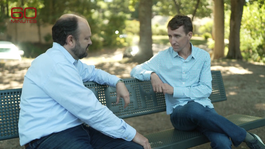 Two men in button-down shirts and jeans sit on a park bench talking.