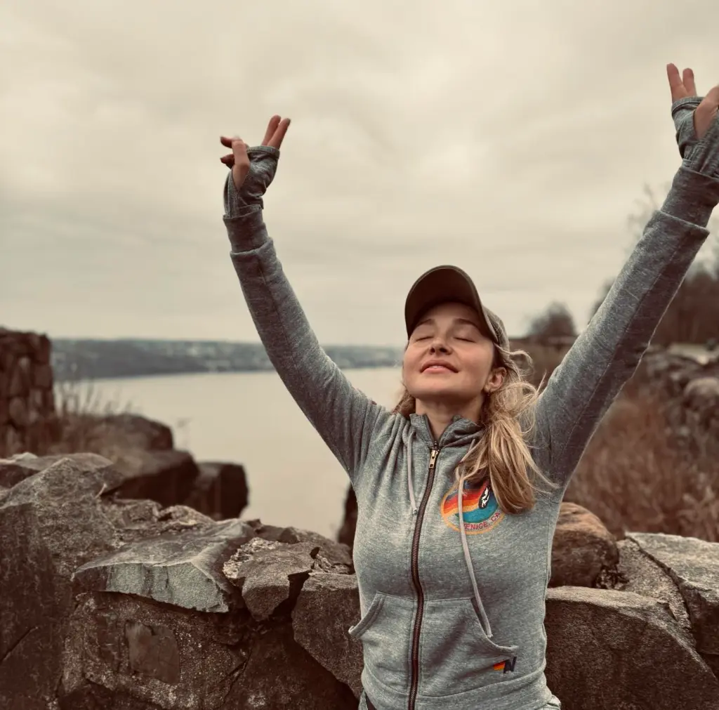 Hayden Panettiere with closed eyes and arms raised on a mountain top.