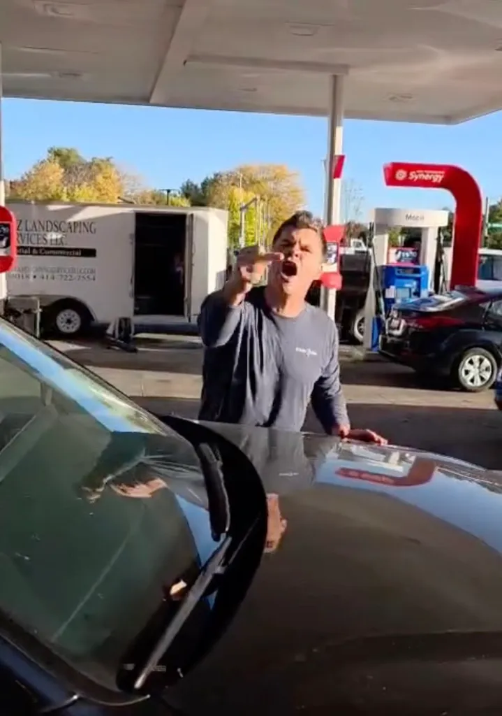 A man gestures and yells angrily next to a car at a gas station.