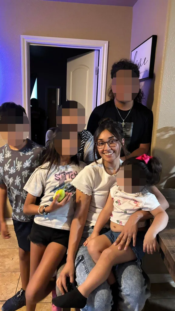 Caroline Peña sits indoors surrounded by several children and a young adult, with the faces of the other individuals blurred out.