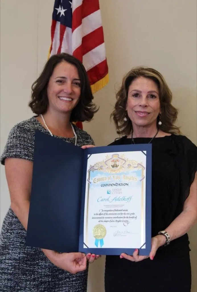 Carol Adelkoff holds a County of Los Angeles commendation certificate alongside another woman, with an American flag in the background.