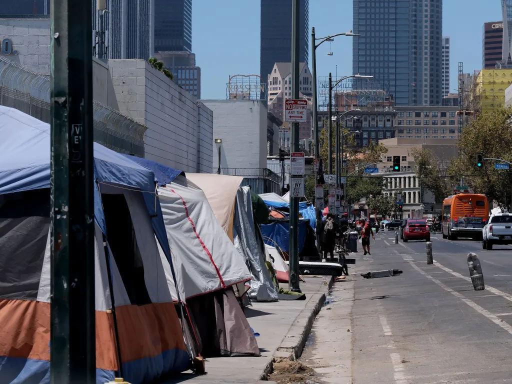 A row of tents lines the sidewalk of a downtown Los Angeles street, with skyscrapers in the background.