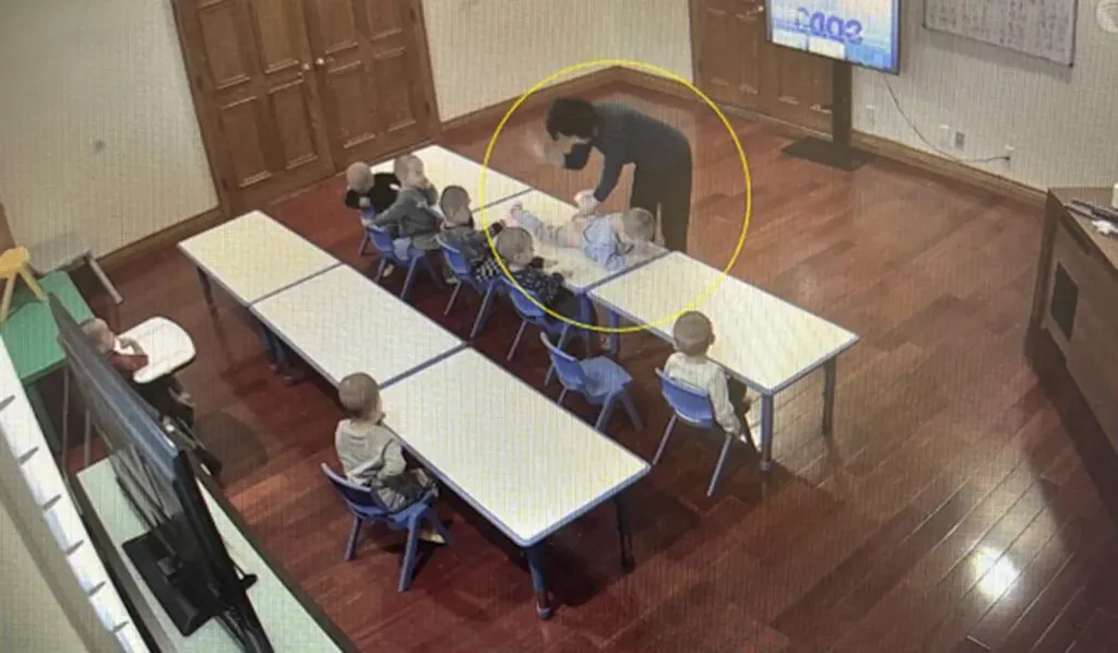 Surveillance footage showing an adult spanking a child lying on a table in a room with other children sitting at tables.