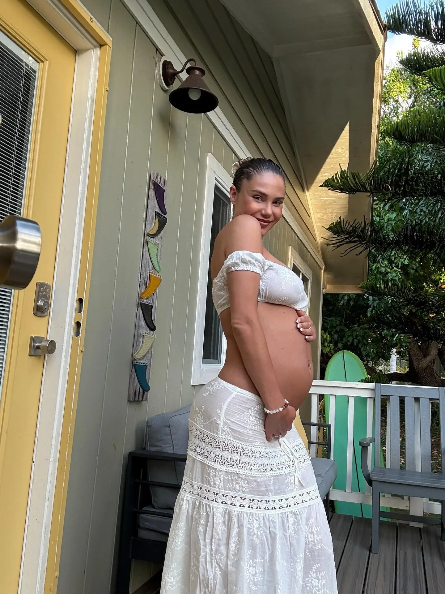 Sahara Ray poses in a white two-piece outfit, cradling her pregnant belly on a porch.