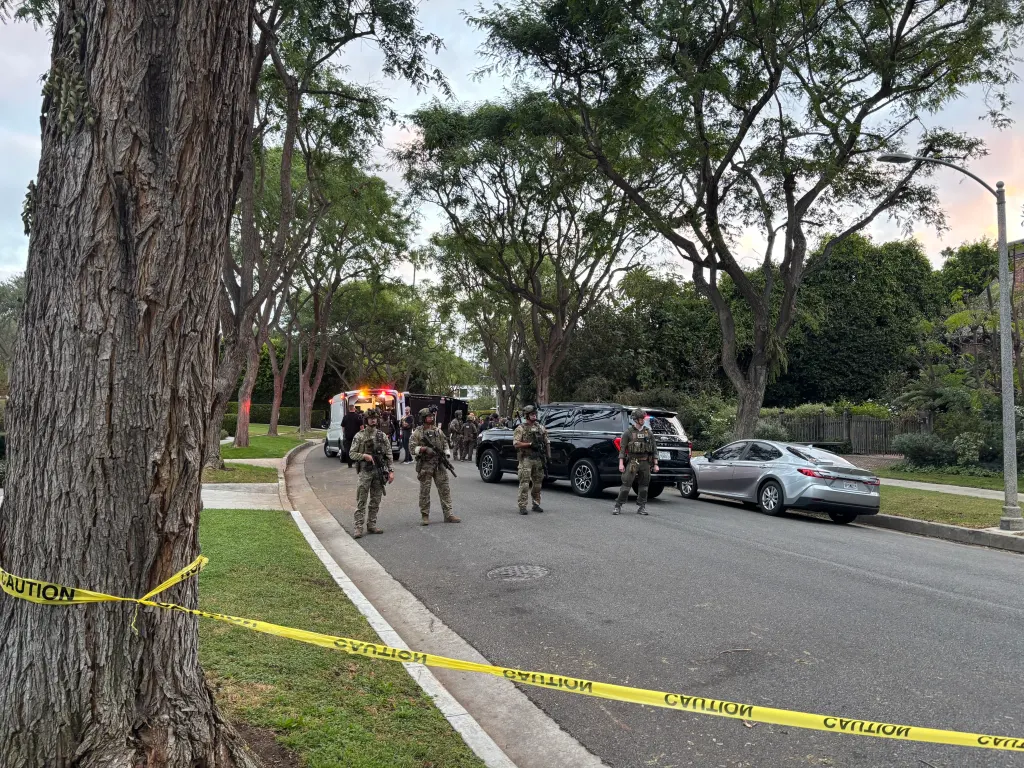 Law enforcement officers in tactical gear stand in a tree-lined residential street behind yellow caution tape, with police vehicles and an ambulance visible in the background.
