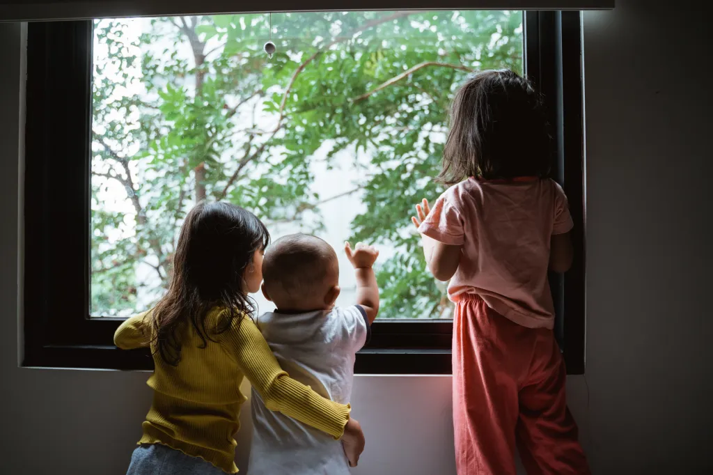 Three young children looking out a bedroom window at green trees.