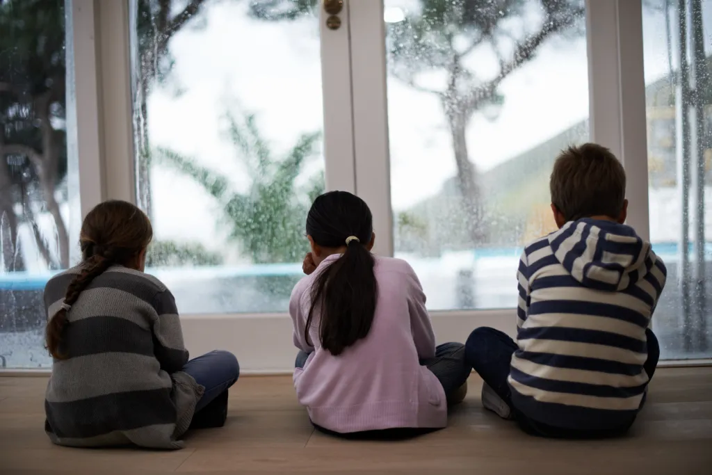 Three children sit on the floor of a living room, looking out a window at the rain.
