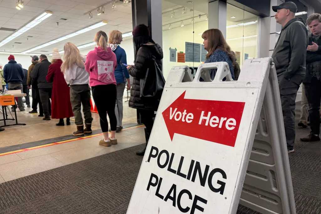 Voters stand in line at an early voting location in a mall in Anchorage, Alaska, with a "Vote Here - Polling Place" sign in the foreground.