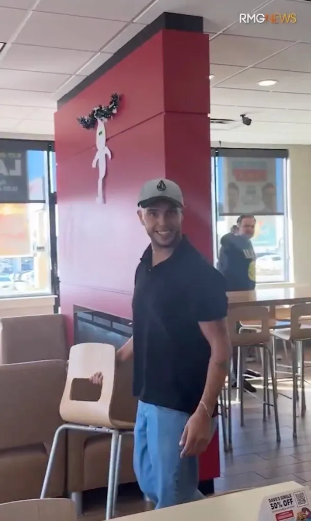 A man in a baseball hat and black polo shirt stands in a fast food restaurant holding a chair during a confrontation.