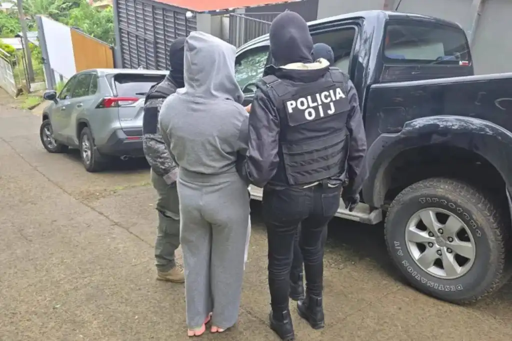 A woman in a grey tracksuit being detained by armed Costa Rican OIJ police officers next to a black pickup truck.